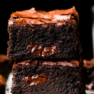 Delicious fudgy brownies stacked on a wooden table, topped with chocolate drizzle.